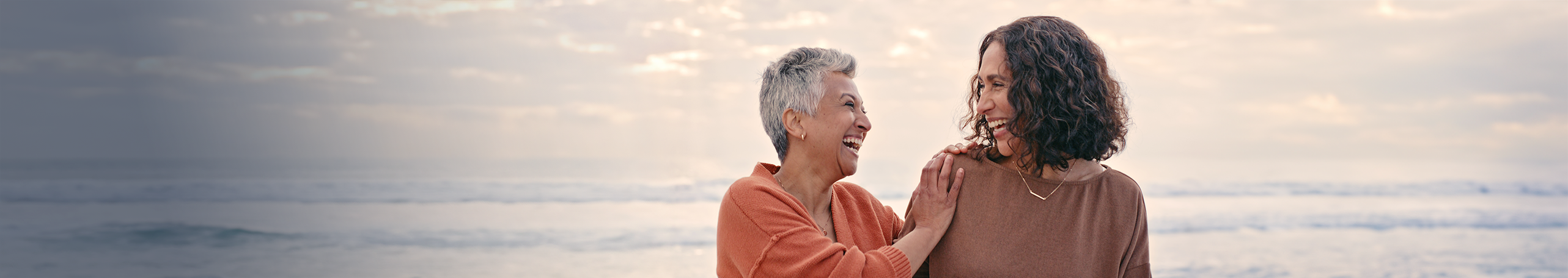 Two diverse women laughing at the beach, possibly discussing a breast screen appointment.